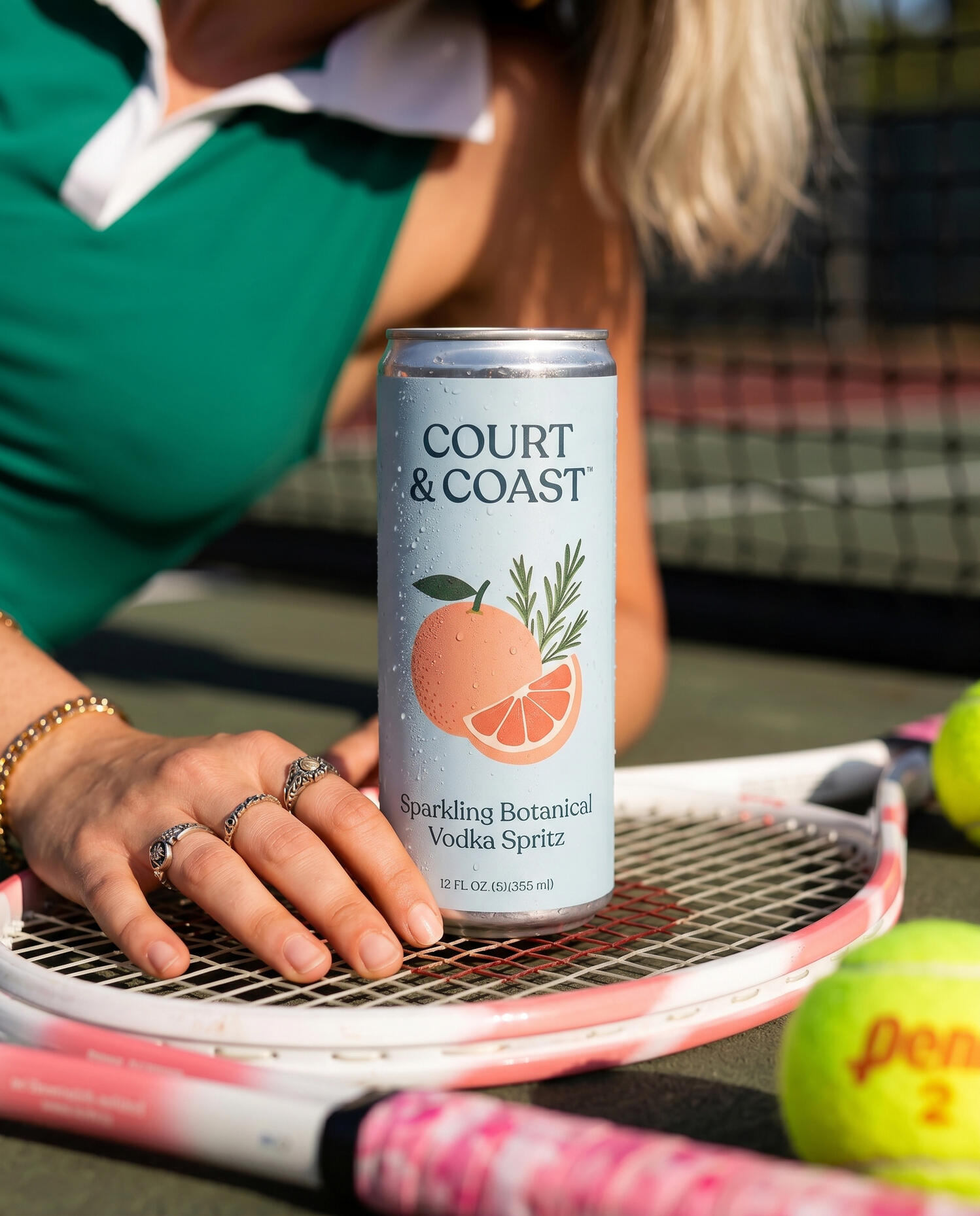 Untitled High-contrast commercial beverage branding mockup, featuring a model in a green athletic outfit holding a canned botanical vodka spritz on a tennis court surface with tennis balls and a racket.