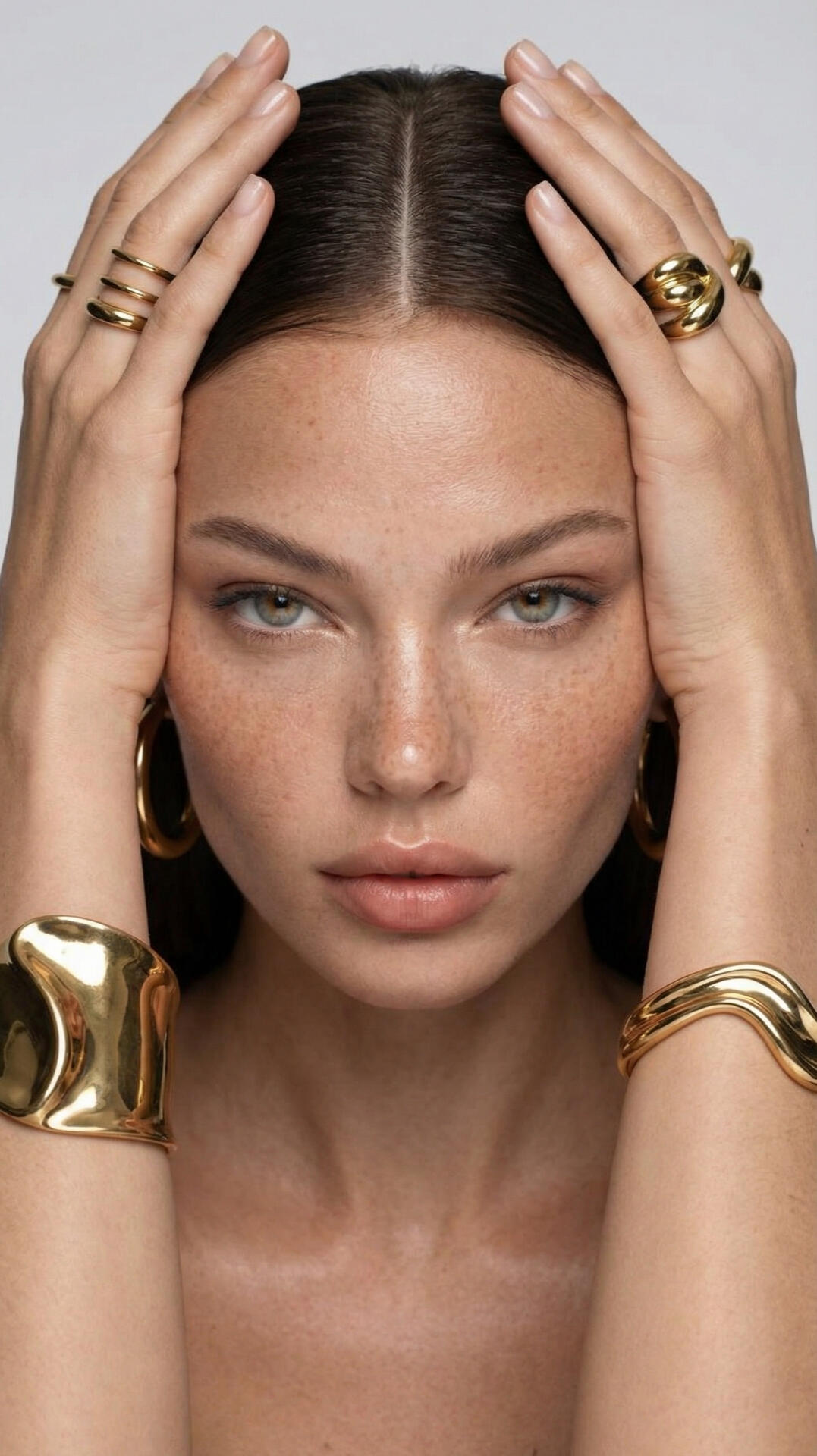 High-fashion jewelry photography and creative direction, featuring a symmetry-framed beauty portrait of a woman styling bold gold statement cuffs and rings.